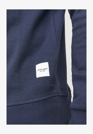 Marineblaue Sweatshirt mit geripptem Saum und Bündchen; verfügt über ein weißes Stoffetikett mit "JACK & JONES EST. 1990" in Schwarz gedruckt.