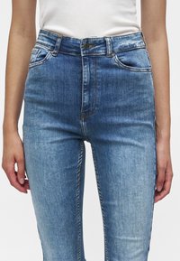 Femme portant un jean bleu clair taille haute avec des poches avant et un haut blanc sans manches, debout avec les bras détendus le long du corps.