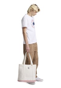 Platnena torba u krem boji s okruglim plavim logom, crvenim naglascima duž dna i dva gornja ručka. Model nosi bijelu majicu i kaki hlače.