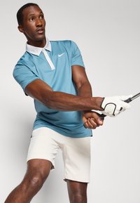 Šviesiai mėlynas Nike polo marškinėliai su balta apykakle, derinami su smėlio spalvos šortais. Modelis laiko golfo lazdą, vilkėdamas baltą pirštinę.