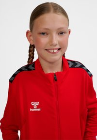 Red zip-up jacket with black accents. Features textured fabric and logo on the chest. Black geometric pattern on shoulders.