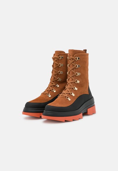 Brown suede boots with black rubber sole and orange accent. Features metallic eyelets and laces. Chunky, lugged sole design.
