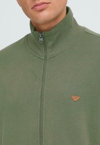 Grüne Zip-Jacke mit hohem Kragen, aus weichem Stoff gefertigt, mit einer strukturierten Innenseite und einem kleinen braunen Logopatch auf der Brust.