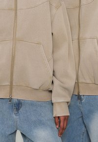 Deux personnes portant des sweats à capuche beige zippés et des jeans bleu clair, debout côte à côte avec une main visible.