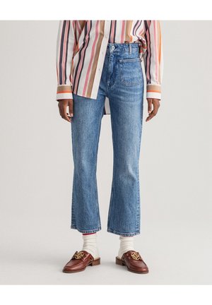 Osoba w krótkich niebieskich dżinsach, paskowanej koszuli na guziki, białych skarpetkach i brązowych mokasynach z złotym łańcuszkiem.