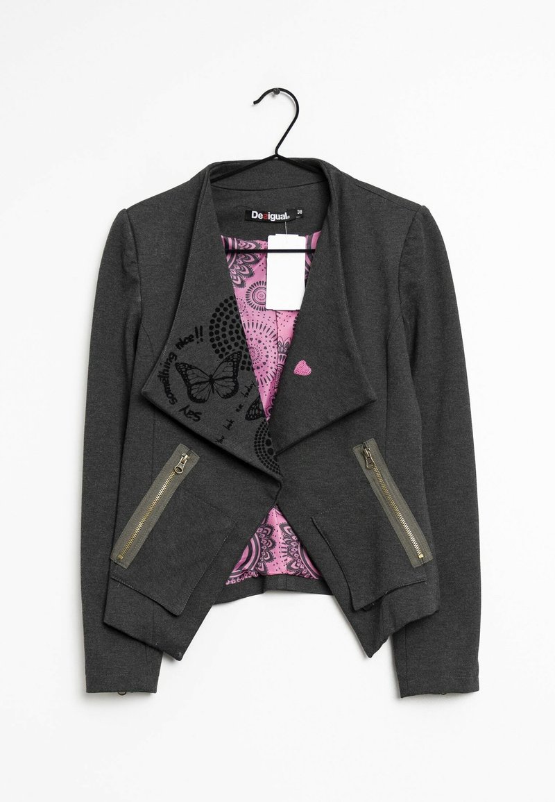 Blazer femme gris foncé avec devant asymétrique, poches zippées, motif papillon à l’intérieur avec doublure rose, suspendu sur un cintre noir.