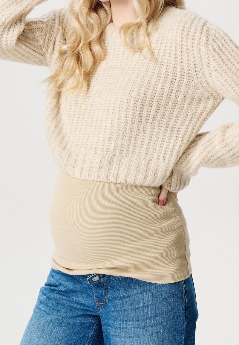 Pull en maille crème avec une texture côtelée, associé à un haut beige ajusté et un jean en denim bleu. La tenue met en valeur des détails de superposition.