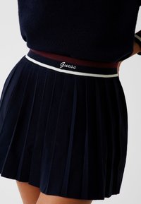 Marineblauer Faltenrock mit einem gerippten Taillenbund, der rot-weiße Streifen und ein Logo aufweist. Der Stoff wirkt strukturiert und hat eine Textur.