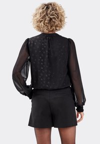 Blouse en chiffon noir avec des manches longues transparentes, arborant un motif scintillant discret et un col froncé, portée avec un short noir.