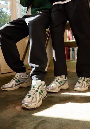 Dwie osoby w srebrno-zielonych butach New Balance, jedna siedząca, druga stojąca, w tle regał z książkami i światło słoneczne.