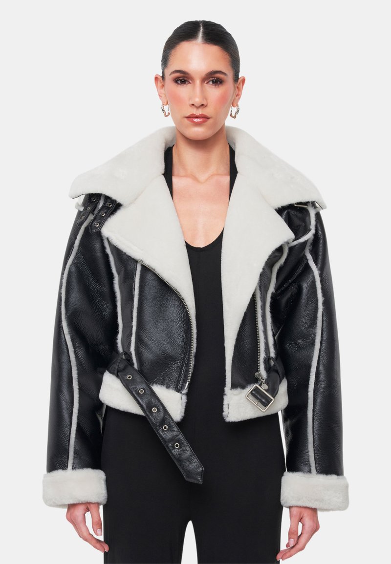 Svart läderjacka med shearling, vit pälskrage, dubbel dragkedja, rem med spänne på sidan och vita sömmar längs sömmarna.