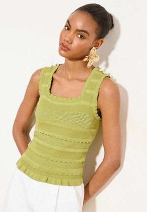 Femme portant un haut tricoté sans manches vert clair avec des motifs texturés et de grandes boucles d’oreilles dorées en forme de fleur sur un fond uni.