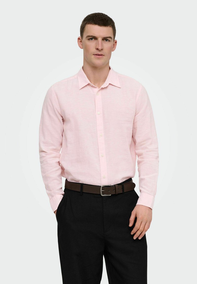 Homme portant une chemise rose clair boutonnée rentrée dans un pantalon noir avec une ceinture marron, debout avec une main dans la poche.