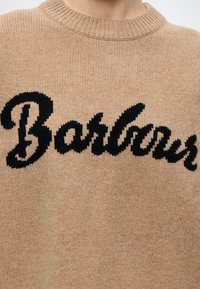 Pull en tricot beige avec texte cursif noir ""Barbour"" sur la poitrine, visible du cou jusqu'au milieu du torse.