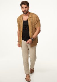 Ein khakifarbendes, offen gestricktes Shirt über einem schwarzen Tanktop, kombiniert mit beigen strukturierten Hosen und braunen Sandalen. Bemerkenswerte Accessoires sind Armbänder und eine Halskette.
