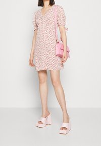 Kvinna klädd i en vit och röd blommig minikjol, rosa platåklackar och bär en liten rosa handväska med axelrem.