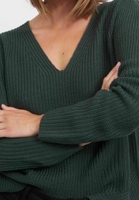 Ciemnozielony sweter dzianinowy z dekoltem w serek, prążkowaną strukturą i długimi rękawami. Podkreśla luźny krój i swobodny wygląd.