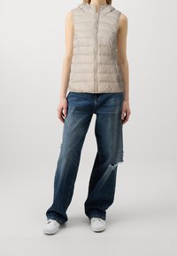 Veste sans manches beige en duvet avec un design matelassé horizontal, une fermeture éclair à l'avant et une capuche. Associée à un jean large bleu déchiré et des baskets blanches.