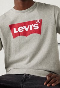 Camiseta de algodón gris con un destacado logotipo rojo de Levi's en un diseño curvado en el pecho. Mangas cortas y cuello redondo acanalado.