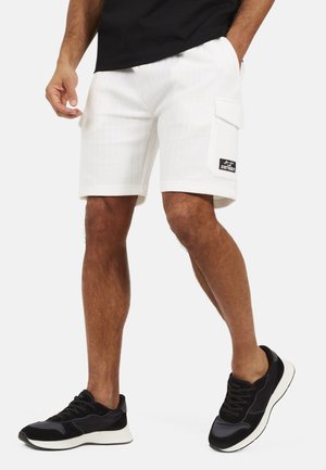 Man draagt witte, tekstuurrijke cargobroek met zijzak en zwarte sneakers, staand met één hand in de zak.