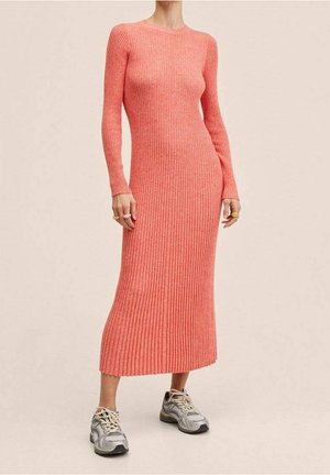 Femme portant une robe midi longue à manches longues en maille côtelée corail ajustée, associée à des baskets grises et blanches, debout sur un fond beige.