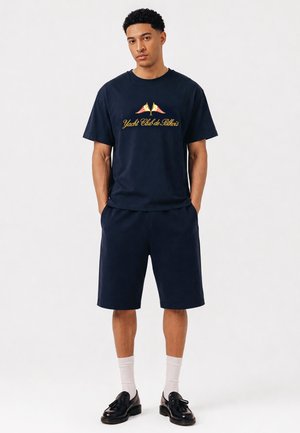 Mann iført marineblå «Yacht Club de Bkirkis» t-skjorte, matchende shorts, hvite sokker og svarte tasselloafers, står med hendene i lommene.