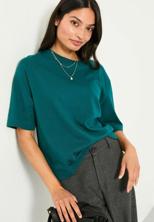 Türkises T-Shirt mit kurzen Ärmeln aus weicher Baumwolle, mit lockerer Passform und rundem Halsausschnitt, kombiniert mit strukturierten, anthrazitgrauen Hosen.