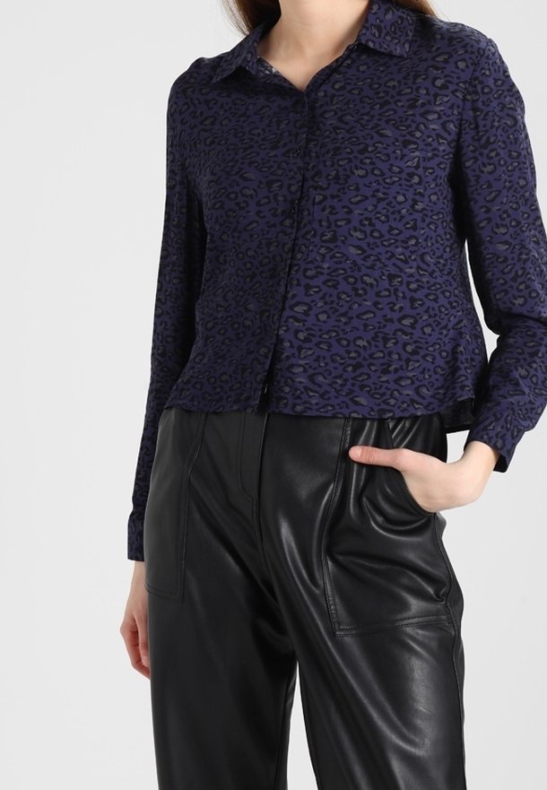 Femme portant une chemise à manches longues avec un imprimé léopard bleu foncé et un pantalon en cuir noir taille haute, une main dans la poche.