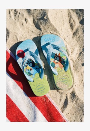 Blauwe kinderslippers met cartoonhondjes erop gedrukt, liggend op zand naast een rood-wit gestreepte strandhanddoek.