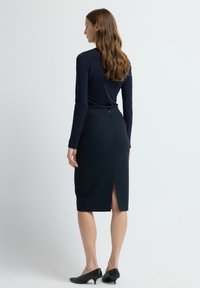 Kobieta z długimi brązowymi włosami ubrana w granatową bluzkę z długim rękawem, czarną spódnicę o długości do kolan z tylnym rozcięciem i niskie czarne obcasy, stojąca bokiem.