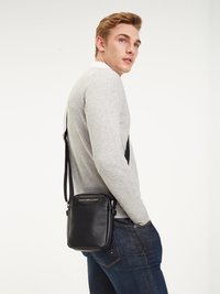 Black leather crossbody bag with adjustable strap, rectangular shape, and embossed logo on the front, worn by a model in a gray sweater.
