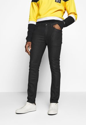 Person wearing slim black jeans, white sneakers, and a yellow and black long-sleeve top with a white waistband, standing with right hand in pocket.