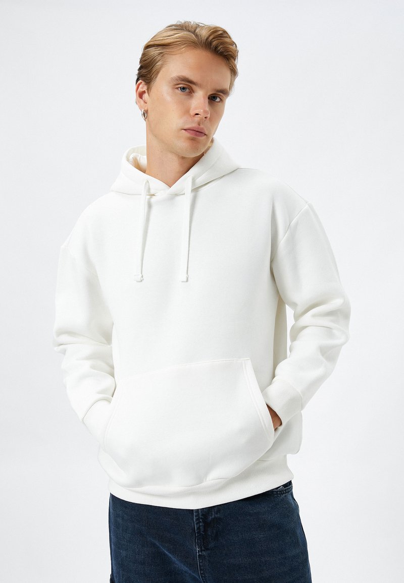 Sudadera blanca hecha de un tejido suave, con un bolsillo frontal, cordones ajustables y puños acanalados. Llevada sobre jeans oscuros.