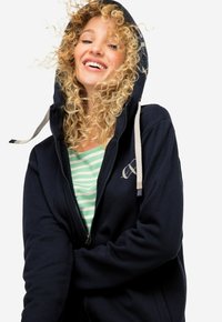 Felpa blue navy con zip, cappuccio regolabile e dettagli con logo. Materiale morbido. Camicia a righe verde chiaro sotto. Capelli ricci visibili.