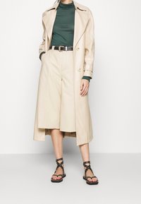 Frau trägt einen beigen langen Trenchcoat, hoch taillierte beigen weit geschnittene Shorts, dunkelgrünen Rollkragenpullover, schwarzen Nieten-Gürtel und schwarze Riemchensandalen.