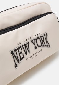 Pochette beige à fermeture éclair avec texte noir "NEW YORK" et texte plus petit "COLLEGE TEAM" et "VARSITY LEAGUE EST. 2001" sur la poche avant.