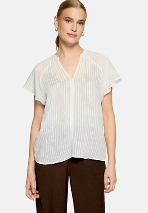 Femme portant un chemisier blanc semi-transparent à manches courtes avec des rayures verticales et un pantalon marron foncé, debout devant un fond uni.