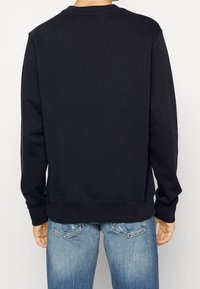 Mężczyzna w prostym czarnym długim rękawie swetrze i jasnoniebieskich dżinsach, pokazany od tyłu na jednolitym tle.