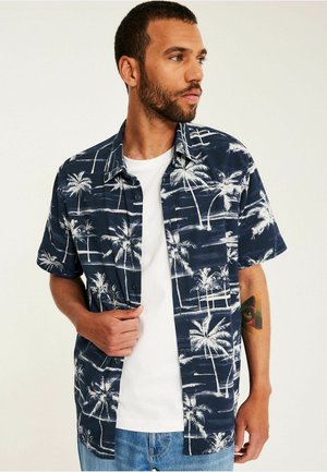 Mann trägt ein dunkelblaues kurzärmeliges Hemd mit weißen Palmenmotiven über einem weißen T-Shirt und blauen Jeans, schaut zur Seite.