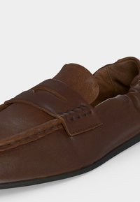 Mocassin marron en cuir pour homme avec détails cousus et semelle en caoutchouc noir, incliné pour montrer l'avant et le côté.