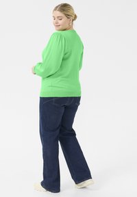 Grøn langærmet top med puffede skuldre, parret med mørkeblå flared jeans. Stoffet ser blødt ud og har en glat tekstur.