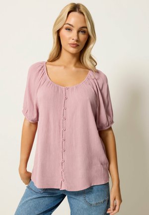 Frau mit blondem Haar, die eine rosa kurzärmelige Bluse mit Knöpfen und blaue Jeans trägt, steht vor einem schlichten hellen Hintergrund.