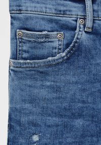 Primer plano de un bolsillo de unos jeans de mezclilla azul con remaches de metal, costura visible, ligero deshilachado y detalle de tela desgastada.
