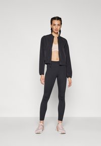 Svart cropped jakke med jevn tekstur, kombinert med en lys rosa sports-bh og høyhårede svarte leggings. Bruker rosa treningssko.