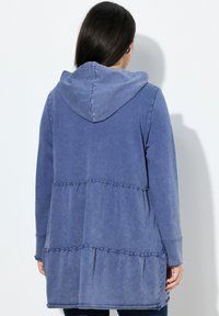Giacca a maniche lunghe con cappuccio in tessuto blu sbiadito, con un design a strati, caratterizzata da cuciture volant e una vestibilità comoda. Materiale testurizzato e morbido.