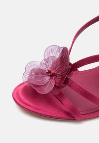 Ροζ σανδάλι ανοιχτής μύτης με λεπτές λουρίδες και διακοσμητικό διάφανο ροζ λουλούδι στο μπροστινό λουρί, τοποθετημένο σε λευκό φόντο.