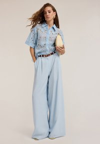 Ljusblå spetsblus med korta ärmar, hög midja och vida byxor i en ljusare blå nyans. Brun bälte och beige handväska, minimala detaljer.