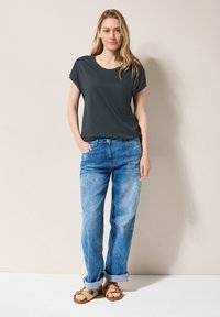 Camisa gris oscuro de manga corta, de corte holgado, combinada con unos vaqueros de pierna ancha en azul claro, ligeramente desgastados, remangados en los tobillos, y sandalias de cuero trenzado marrones.