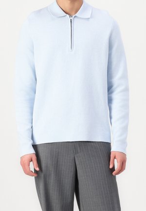 Mann trägt hellblaues Langarm-Poloshirt mit Frontreißverschluss und graue Nadelstreifenhose, steht vor weißem Hintergrund.