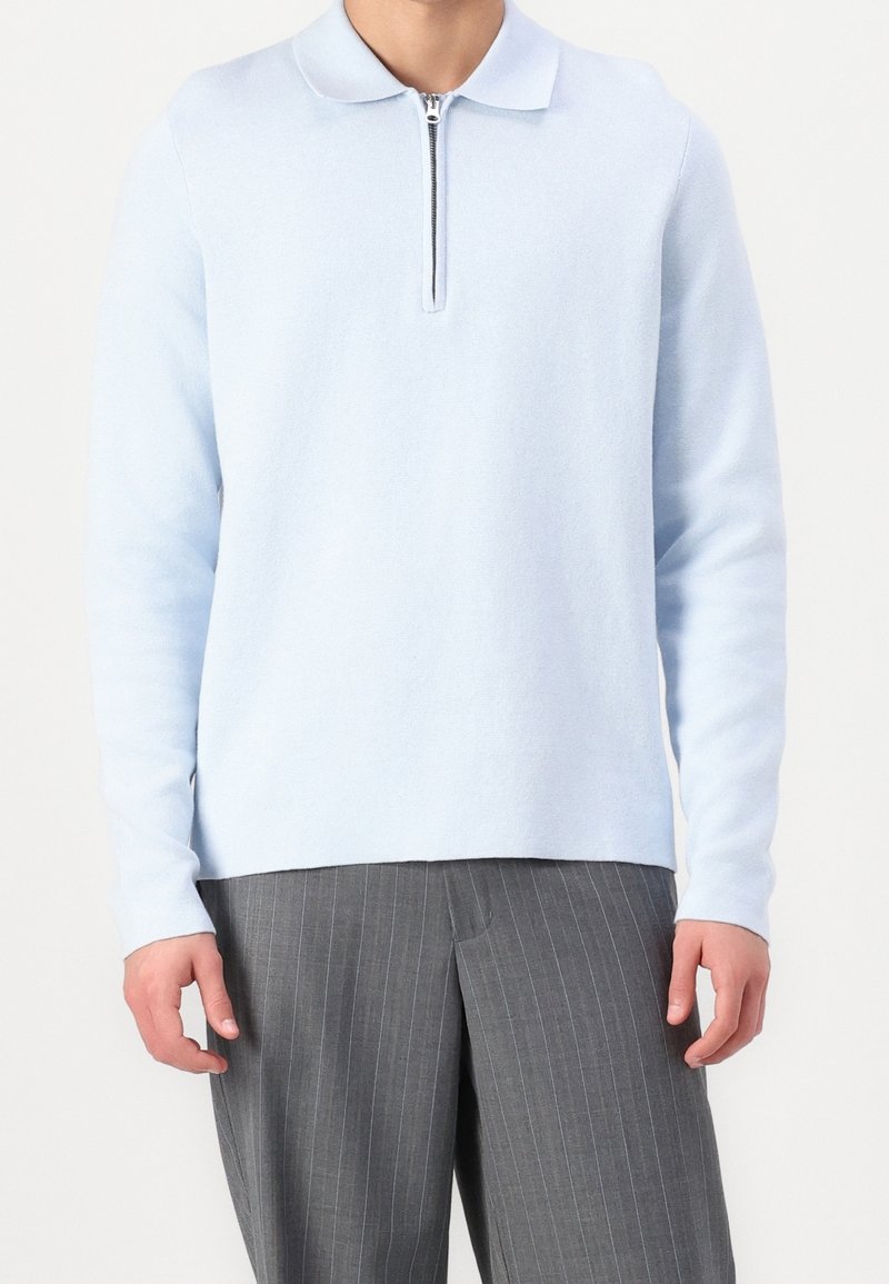 Homme portant un pull polo à manches longues bleu clair avec fermeture éclair sur le devant et un pantalon gris à fines rayures, debout devant un fond blanc.